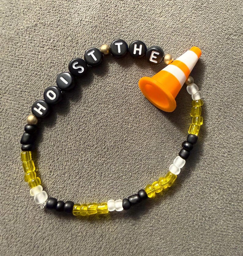 May include: A beaded bracelet with the words "HOIST THE" spelled out in black letter beads. The bracelet features a small orange and white traffic cone charm, and is strung with yellow, black, and white beads.