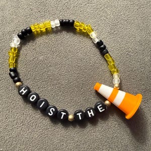 May include: A beaded bracelet with the words "HOIST THE" spelled out in black letter beads. The bracelet features yellow, black, and white beads, and an orange and white traffic cone charm.