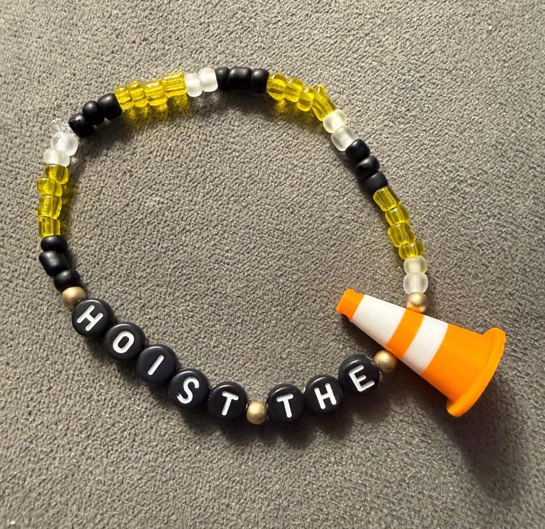 May include: A beaded bracelet with the words "HOIST THE" spelled out in black letter beads. The bracelet features yellow, black, and white beads, and an orange and white traffic cone charm.
