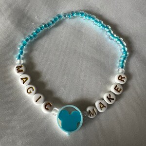 May include: A blue and white beaded bracelet with the words "MAGIC MAKER" spelled out in white beads with gold lettering. A blue Mickey Mouse head charm is attached to the bracelet.