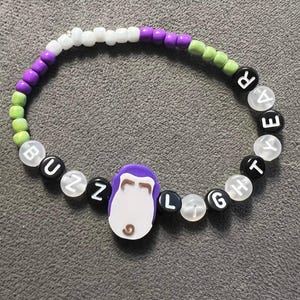 May include: A purple, white, green and black beaded bracelet with the words "Buzz Lightyear" spelled out on it.