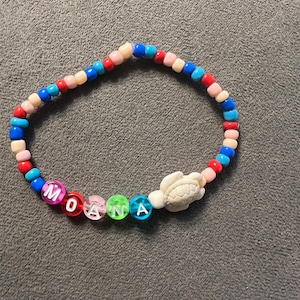 Könnte beinhalten: Ein farbenfrohes Perlenarmband mit dem Namen "MOANA", der in Buchstabenperlen geschrieben ist. Das Armband hat außerdem einen weißen Schildkrötenanhänger.