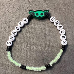 May include: A green and white beaded bracelet with the words "SNACKY" and "SNACKS" spelled out on it. The bracelet also features a green character charm.
