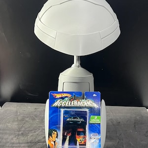 Op de afbeelding: Een witte, 3D-geprinte displaystandaard met een Hot Wheels Acceleracers speelgoedautopakket. Het pakket bevat een blauwe en oranje speelgoedauto, met de tekst "Acceleracers" en "3+". De standaard heeft een futuristisch, helmachtig ontwerp.