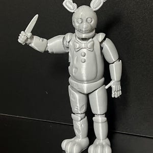 Pode incluir: Uma figura cinzenta articulada de um personagem semelhante a um coelho, segurando uma faca. A figura tem uma gravata borboleta, detalhes de botões e orelhas de coelho. A figura está em pé sobre uma superfície preta.