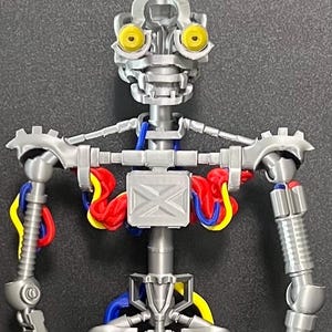 Pode incluir: Um brinquedo de esqueleto de robô articulado prateado com olhos amarelos, fiação azul e amarela e detalhes em vermelho. O brinquedo tem um design mecânico detalhado, com juntas e engrenagens visíveis, e está em posição vertical.