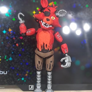 Könnte beinhalten: Ein rotes und graues animatronisches Fuchsspielzeug mit einem Haken als Hand, einem Augenklapp und einem fehlenden Kiefer. Das Spielzeug steht auf einer Holzoberfläche.