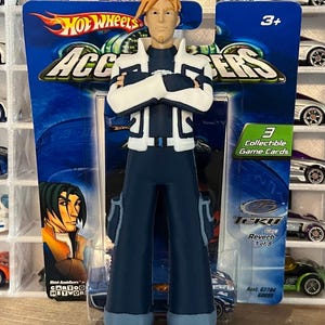 Op de afbeelding: Een blauwe en witte action figure van een personage uit de animatieserie 'Hot Wheels AcceleRacers'. De figuur heeft zijn armen gekruist en heeft een vastberaden uitdrukking op zijn gezicht. De figuur is verpakt in een blauwe en groene kartonnen doos met het Hot Wheels logo en de woorden 'AcceleRacers' erop gedrukt. De doos bevat ook een klein kaartje met de woorden 'Teku' en 'Reverb 1 of 9' erop gedrukt.