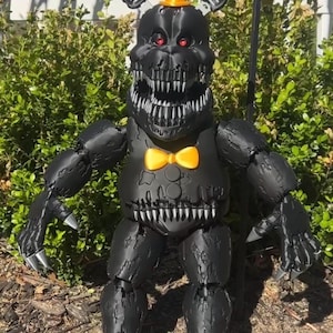 Pode incluir: Uma estatueta preta de uma criatura parecida com um urso de pesadelo com dentes afiados, olhos vermelhos e uma gravata borboleta amarela. Possui uma cartola e mãos em forma de garra. A figura está ao ar livre em frente a arbustos verdes e uma cerca branca.