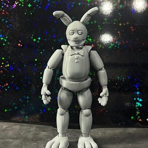 May include: A gray articulated rabbit figure with a bow tie, long ears, and large feet. The figure is standing on a gray surface with a dark background featuring colorful star-shaped patterns. The figure has a smiling expression.
