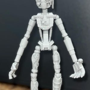 May include: A gray 3D printed articulated robot figure with a detailed head and body. The robot has a human-like form with a long neck, long arms, and long legs. The robot's hands and feet are articulated.
