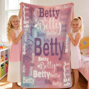 May include: A personalized blanket in shades of pink, purple, and blue, featuring the name "Betty" in various fonts and sizes. The blanket is held up by two young girls in a well-lit room, showcasing the custom design.