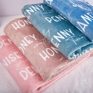 May include: A stack of soft blankets in shades of pink, peach, and blue. Each blanket features white text with the words "HONEY", "PENNY", and "AMY". The blankets are folded and stacked on top of each other.