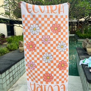May include: A white and orange checkered beach towel with the name "ELVIRA" printed in orange. The towel features daisy flower graphics in pink, white, and orange. The towel is held up in front of a pool.