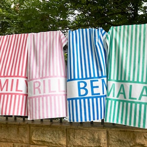 May include: Four striped beach towels in pink, blue, and green, each with a white band featuring a name in bold letters: "EMILY", "RILL", "BEMALA", and "BEMALA". The towels are hanging on a wrought iron fence.
