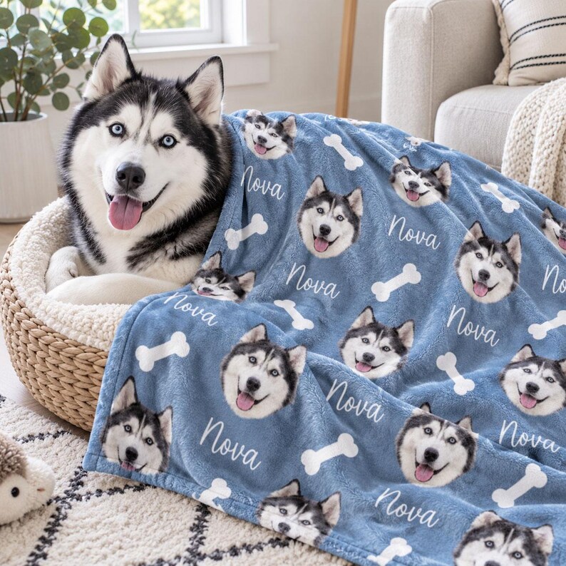 May include: A blue fleece blanket featuring a repeating pattern of a Siberian Husky dog's face, the name Nova in white script, and white bone shapes. The blanket is draped over a dog resting in a woven pet bed.