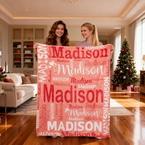 Könnte beinhalten: Eine korallenfarbene Decke mit dem Namen "Madison" in Rot und Weiß mehrfach bedruckt. Zwei Personen halten die Decke in einem hellen Raum, mit einem Weihnachtsbaum im Hintergrund. Das Design beinhaltet Herzen und verschiedene Schriftarten.