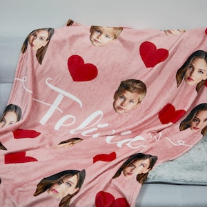 May include: A pink fleece blanket with multiple faces and red hearts. The blanket has the name "Felicia" in white script. The blanket is draped over a grey sofa and a fluffy white rug.