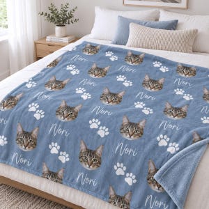 May include: A blue fleece blanket featuring a repeating pattern of a tabby cat's face, white paw prints, and the name Nori written in a white cursive script. The blanket is draped over a bed with white pillows and a blue accent pillow.