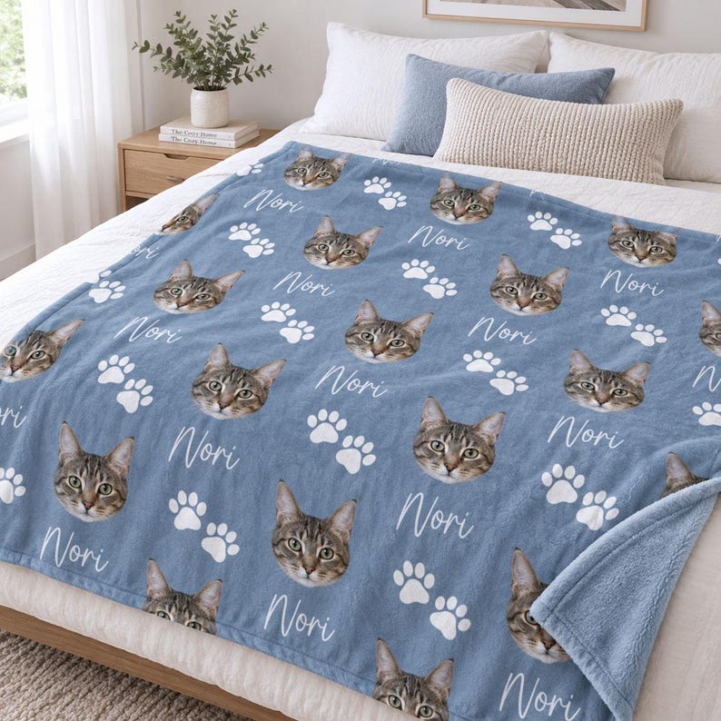 May include: A blue fleece blanket featuring a repeating pattern of a tabby cat's face, white paw prints, and the name Nori written in a white cursive script. The blanket is draped over a bed with white pillows and a blue accent pillow.