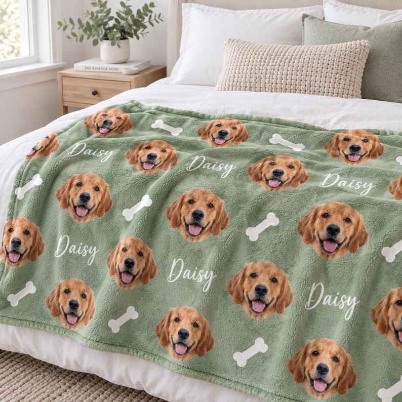 May include: A sage green fleece blanket featuring a repeating pattern of golden retriever dog faces and white bone shapes. The name Daisy is printed in a white cursive script next to each dog face across the entire surface of the soft fabric.