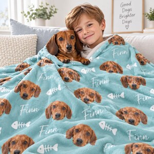 May include: A light blue fleece blanket featuring a repeating pattern of a brown dachshund dog face, the name Finn in white script, and white fish bone icons. The soft fabric displays multiple instances of the dog's portrait and the name.