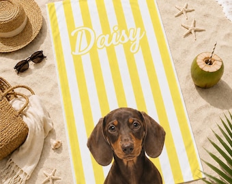 May include: A yellow and white striped beach towel with the name "Daisy" in script. The towel features a brown dachshund dog at the bottom. The scene includes a straw hat, sunglasses, a tote bag, and a coconut on a sandy beach.