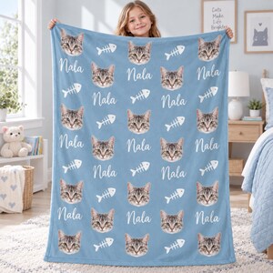 May include: A light blue fleece blanket featuring a repeating pattern of a grey tabby cat face, white fish bone skeletons, and the name Nala written in a white cursive script. The design is printed across the entire surface of the fabric.