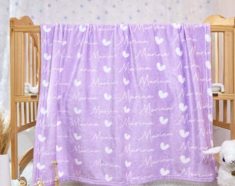 May include: A lavender blanket with the name "Marina" in white script, interspersed with white heart shapes. The blanket is draped over a wooden crib, creating a cozy and personalized nursery item.