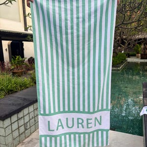 May include: A large, vertically striped beach towel in white and mint green. The word "LAUREN" is printed in mint green on a white band at the bottom of the towel. The towel is held up in front of a pool.
