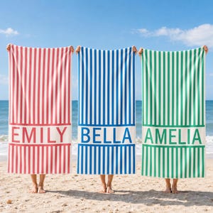 Op de afbeelding: Drie rechthoekige strandlakens met verticale strepen in rood, blauw en groen. Elk laken heeft een witte horizontale band aan de onderkant met de namen EMILY, BELLA en AMELIA in dikgedrukte, hoofdletters gedrukt op de witte strook.