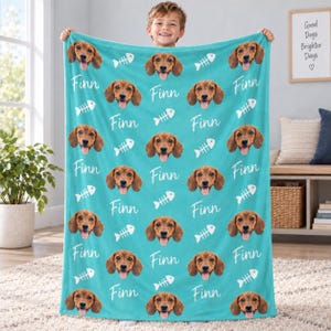 May include: A teal fleece blanket featuring a repeating pattern of a brown dachshund dog face, the name Finn in white script, and white fish bone icons. The soft fabric displays the dog's smiling expression and floppy ears across the entire surface.