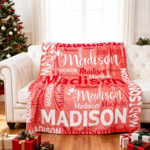 May include: A red blanket featuring the name "Madison" in white and red lettering. The blanket is displayed on a white sofa, with a Christmas tree and wrapped gifts visible in the background.