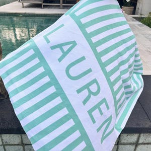 May include: White and mint green striped beach towel with the name "LAUREN" printed in large mint green letters. The towel is folded and draped over a dark surface, with a pool in the background.