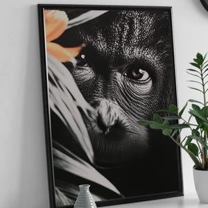 May include: Framed black and white print featuring a close-up of an orangutan's face, partially obscured by foliage and a single orange flower. The print is in a black frame, displayed on a white shelf with a small vase and potted plant.