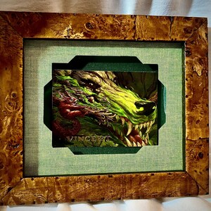 May include: Framed artwork featuring a detailed illustration of a snarling wolf's head with sharp teeth, rendered in shades of green and yellow. A small red creature is on the wolf's face. The artwork is set within a decorative wooden frame.