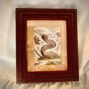 May include: A framed artwork featuring a detailed illustration of a serpentine creature with a red tongue and smoke-like breath. The artwork is set against a beige background and framed in a dark brown, textured wooden frame.