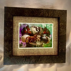 May include: Framed artwork featuring a badger warrior holding a skeleton. The artwork has a fantasy theme with a color palette of brown, white, red, green, and purple. The frame is dark with a textured appearance, and the artwork is set against a neutral background.
