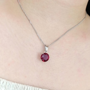 May include: A silver necklace with a round, deep red pendant. The pendant is the focal point, suspended from a delicate chain. The necklace is worn against a light-colored top.