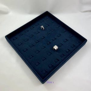 May include: A square, dark blue velvet jewelry display tray with multiple slots for rings. Two silver-toned rings are placed on the tray, one featuring a thin band and the other a wider, multi-layered design. The text Size 1 is visible.