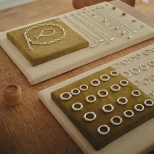 Puede incluir: Una colección de joyas de plata expuestas sobre dos almohadillas de terciopelo verde oliva y una base beige. El conjunto incluye un collar de cadena de plata, una pulsera a juego, varias cadenas finas y numerosos anillos de plata.