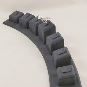 May include: A dark gray velvet jewelry display stand featuring a curved base with six individual square pedestals, each with a small slot. A silver-toned ring with a white pearl and small clear stones is placed on the fourth pedestal from the left.