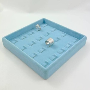 May include: A square light blue velvet jewelry display tray featuring multiple slots for rings. The tray contains two silver rings, one thin band and one wider textured band, positioned within the organized grid of the soft blue display surface.