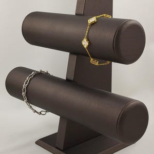 以下が含まれることがあります： ダークブラウンの2段式円筒形ジュエリーディスプレイスタンド。上段には白いクローバー型のチャームが付いたゴールドトーンのチェーンブレスレットが掛けられ、下段にはシルバートーンのチェーンブレスレットが掛けられています。