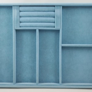 Puede incluir: Una bandeja organizadora de joyas rectangular de color azul claro con un acabado suave similar al terciopelo. El interior está dividido en varias secciones, incluyendo una fila de cuatro ranuras acolchadas para anillos y varios compartimentos.