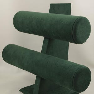 May include: A dark green jewellery display stand with two horizontal, cylindrical bars. The stand is made of a soft, velvety material, designed to hold bracelets or necklaces. The base is square, and the stand is on a white background.