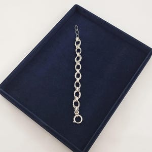 May include: A silver-toned chain bracelet featuring a series of alternating circular links and twisted rope-textured oval links. The bracelet includes a small circular spring ring clasp at one end and a series of smaller adjustment links at the other.