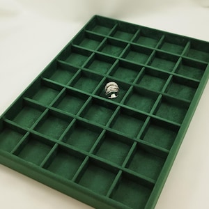 Op de afbeelding: Een groene fluwelen ringen display met 36 vakjes. De display is leeg behalve één vakje dat een zilveren ring bevat.