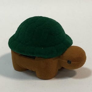 May include: A small, green and brown turtle-shaped ring box. The box is made of velvet and has a hinged lid.