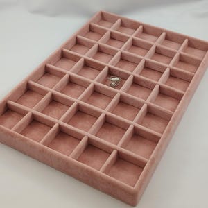 Puede incluir: Una bandeja rectangular de joyería de terciopelo rosa empolvado con múltiples compartimentos cuadrados. Un compartimento contiene un anillo de plata. La bandeja está diseñada para organizar y exhibir joyas, como anillos, pendientes y collares.